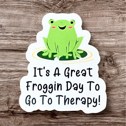A colorful vinyl sticker featuring a smiling green frog sitting on a yellow circle with the phrase 'It's a GREAT FROGGIN DAY TO GO TO THERAPY!' in bold, cheerful lettering.